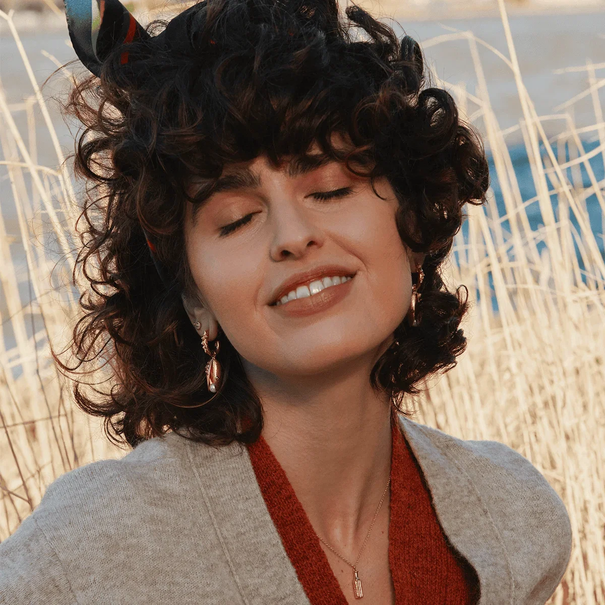 Model wearing Alex Monroe Statement Mussel & Pearl Drop Earrings from Waters Edge Collection, stood amongst coastal grasses.