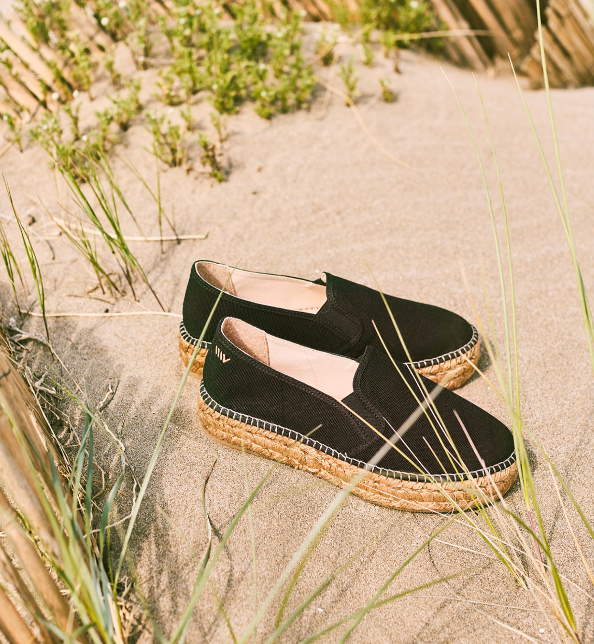 Black canvas platform espadrilles with braided jute soles resting in sunlit sand and beach grass for coastal days