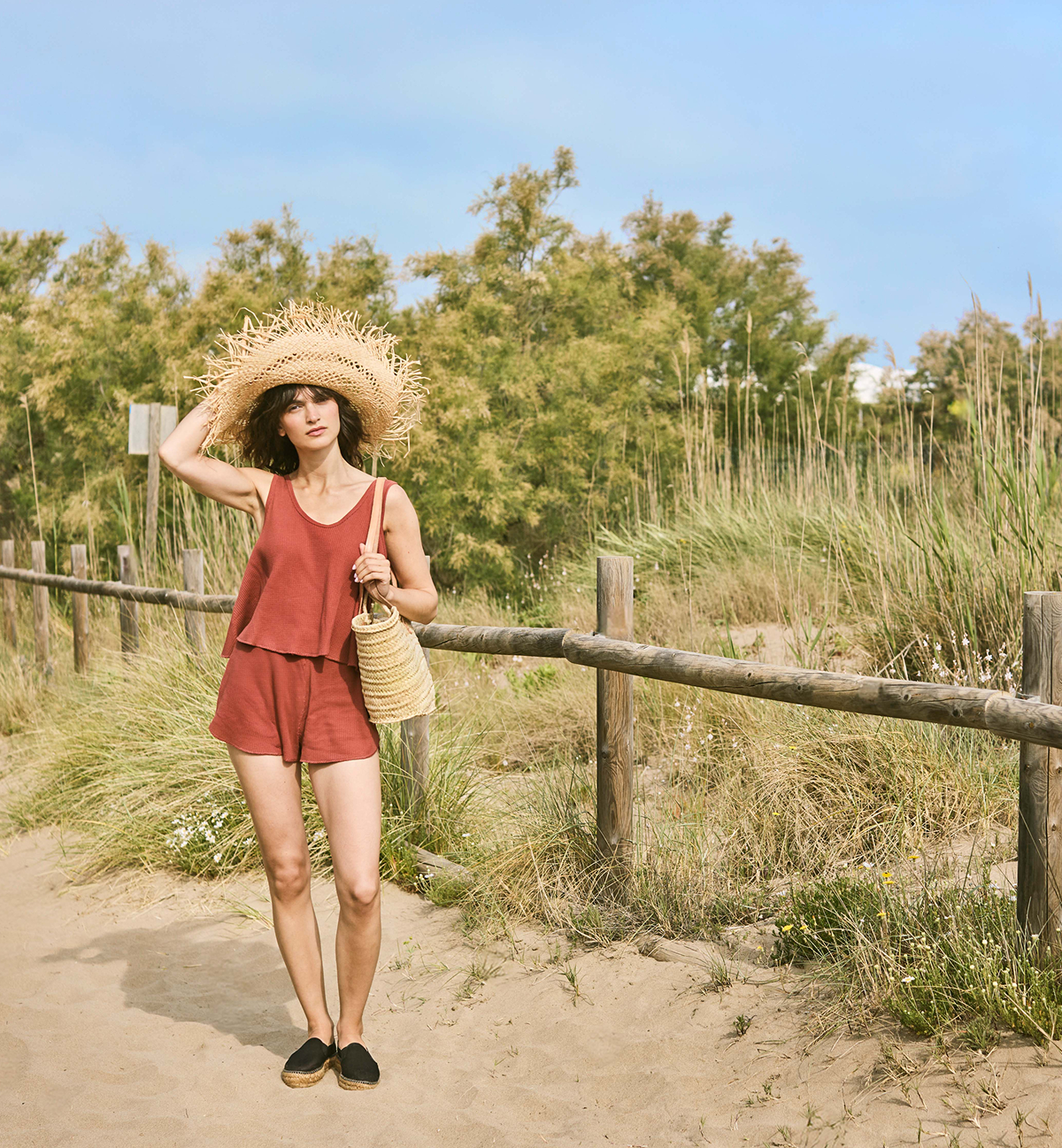 Woman wearing black espadrille flatforms, a rust-colored summer set, and a straw hat while walking through coastal dunes.