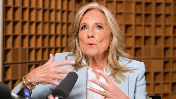  Jill Biden looks upward as she speaks into a mic. She wears a light blue blazer and a white shirt. 