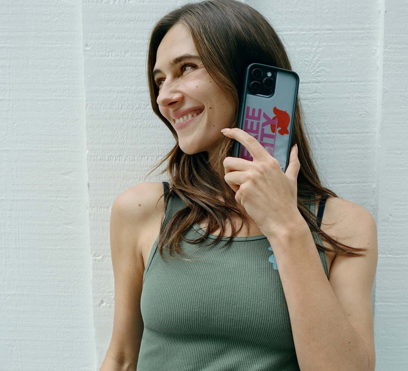 Devon Carlson smiling while holding a with a FREECITY x Wildflower phone case against the side of her face, wearing a ribbed olive green tank top and light denim jeans, standing against a white textured wall outdoors.