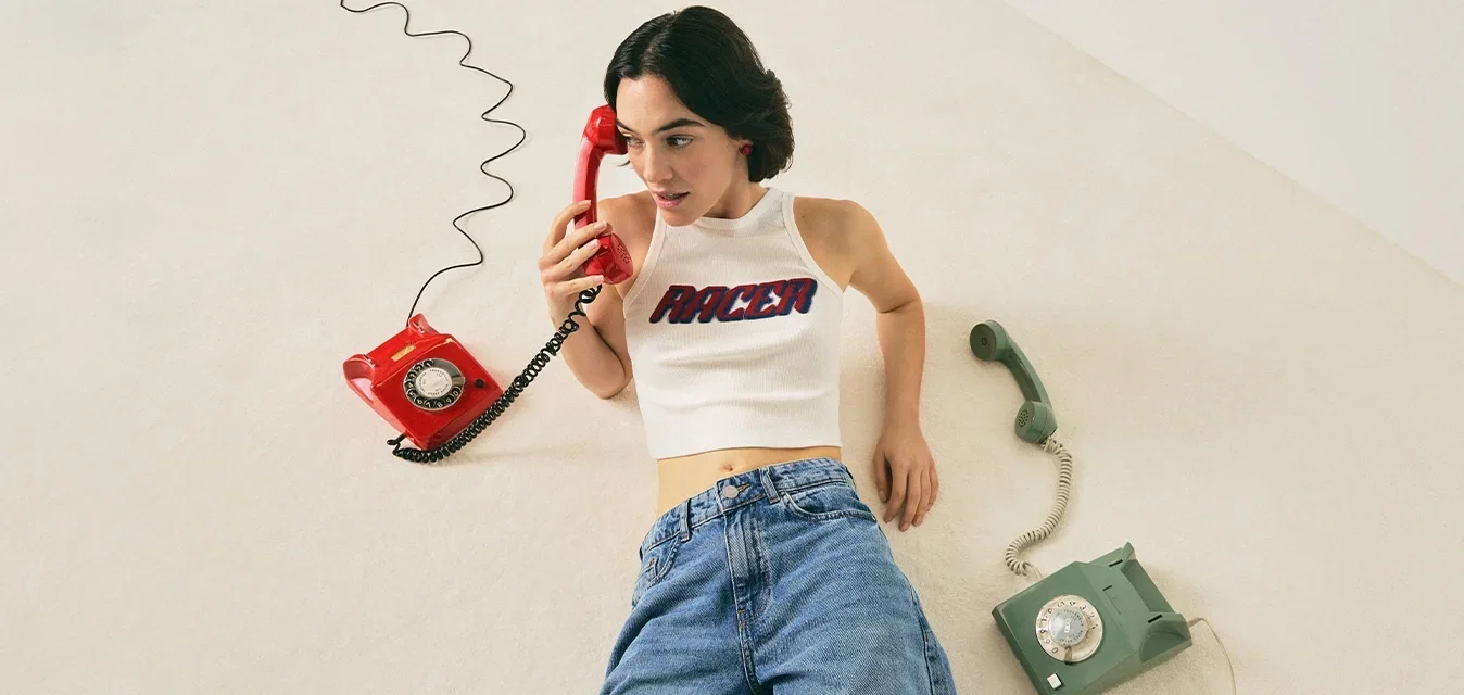 Frau in weißem Crop-Top und Jeans, die auf dem Boden liegt und ein rotes Wählscheibentelefon hält, mit einem grünen Wählscheibentelefon neben sich.