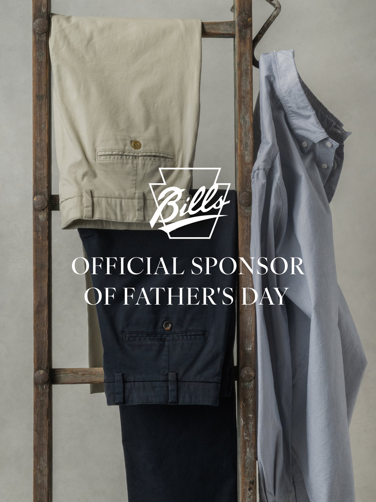 Two pairs of Bills Khakis pants and a Bills Khakis Standard fit shirt hang from a vintage ladder. Text overlay has the Bills Khakis logo and text reads 'Official Sponsor of Father's Day'