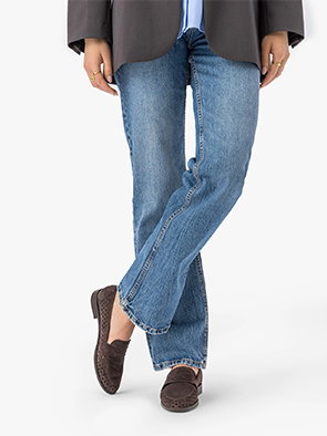 Person wearing blue jeans and brown loafers, standing with legs crossed.
