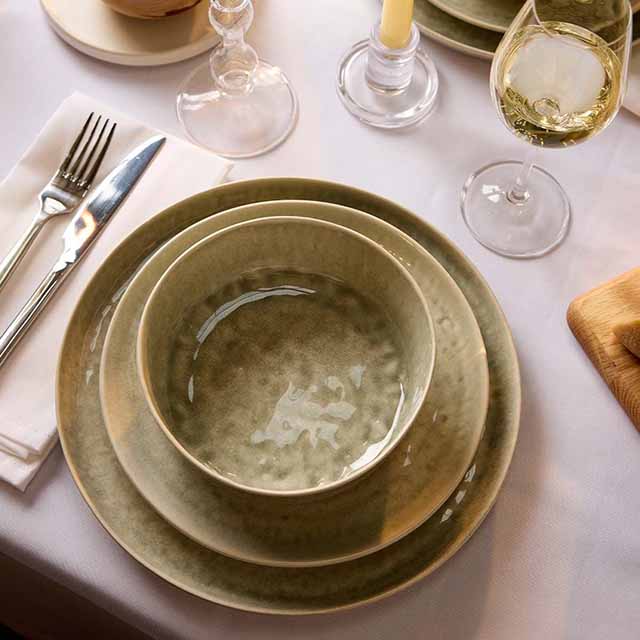 A table setting with beige ceramic dishes, a wine glass, candle, and cutlery on a white tablecloth.