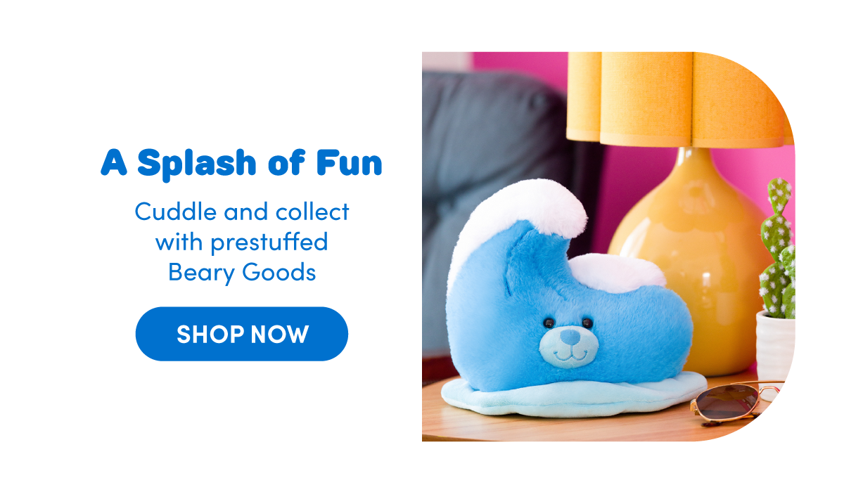 Image featuring a blue wave-shaped plush with a smiling face and white crest detail resting on a light blue base, placed on a tabletop beside sunglasses, a yellow lamp, and small decor against a pink wall backdrop. | Text reads: A Splash of Fun Cuddle and collect with prestuffed Beary Goods | SHOP NOW