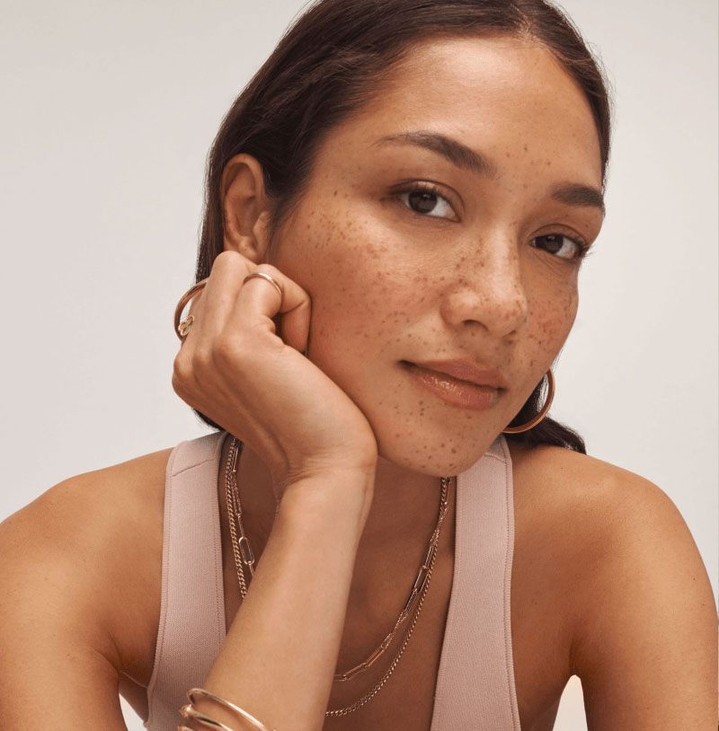 Close-up of female model wearing layered yellow gold chain necklaces, hoop earrings, bracelet, and dainty ring.