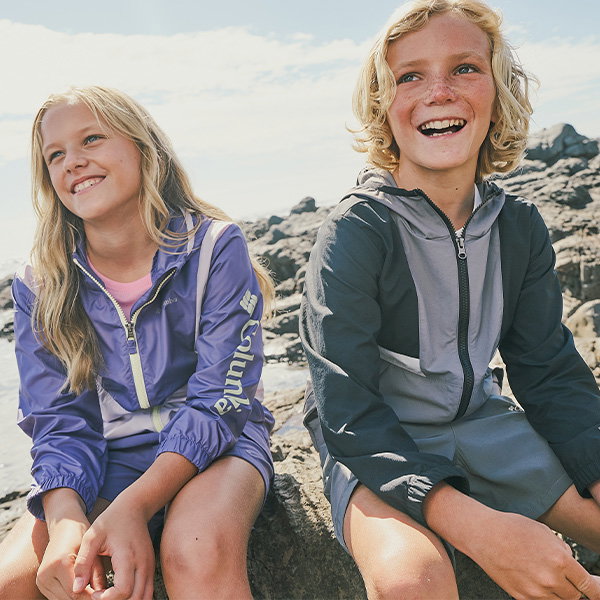 Two children in lightweight jackets sit on sunlit rocks by the coast, with the sea and shoreline stretching behind them.