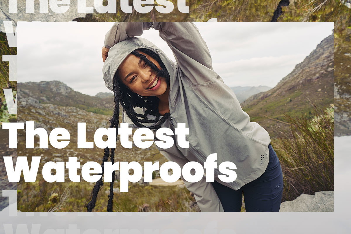 A person in a light grey hooded waterproof jacket leans sideways in a rocky, mountainous landscape with grasses and overcast skies, while large white text across the image reads “The Latest Waterproofs.”