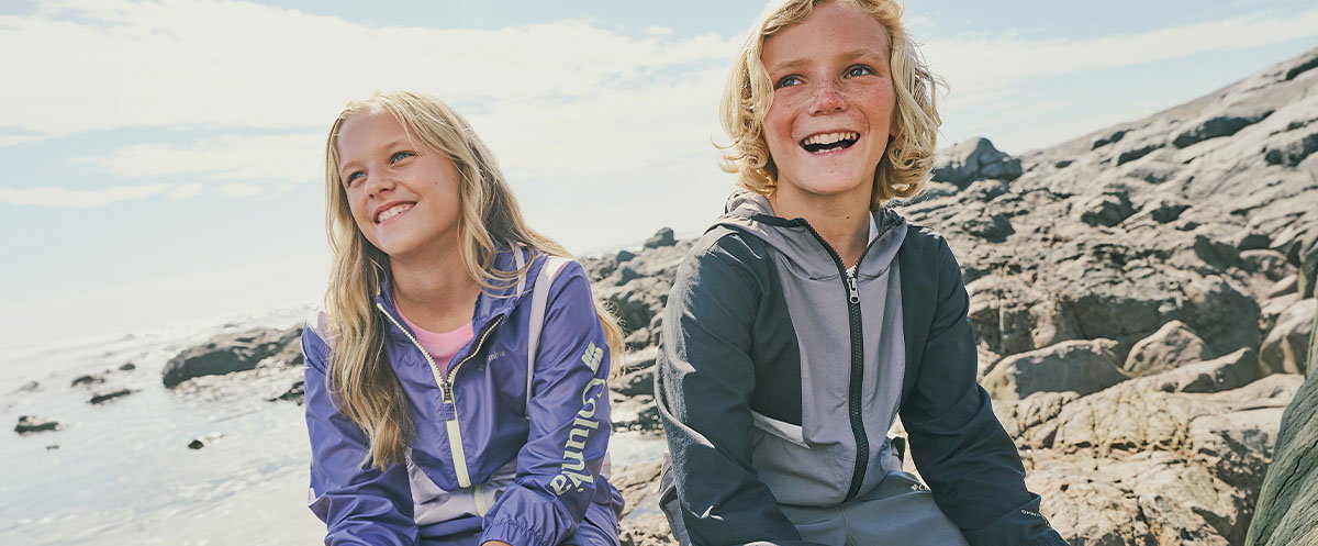 Two children in lightweight jackets sit on sunlit rocks by the coast, with the sea and shoreline stretching behind them.