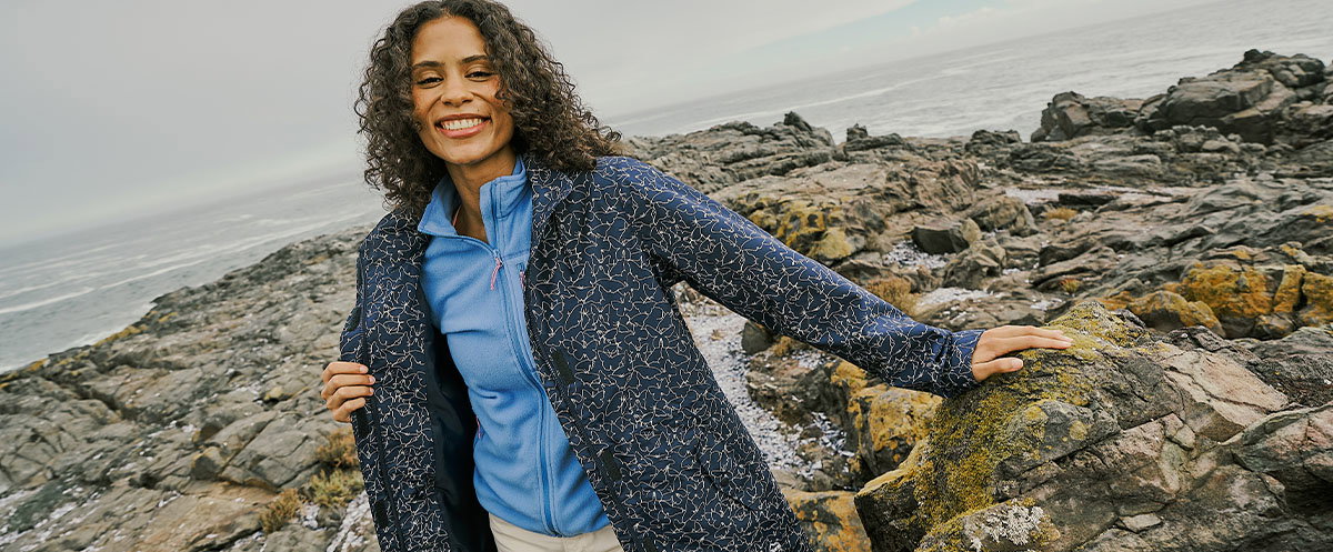A person in a patterned jacket stands on a rocky coastline, holding the jacket open to reveal a blue layer underneath, with the sea and rugged rocks in the background.