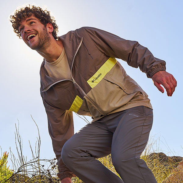 A person in a lightweight brown jacket with a bright panel leans forward outdoors under a clear sky, surrounded by low vegetation.