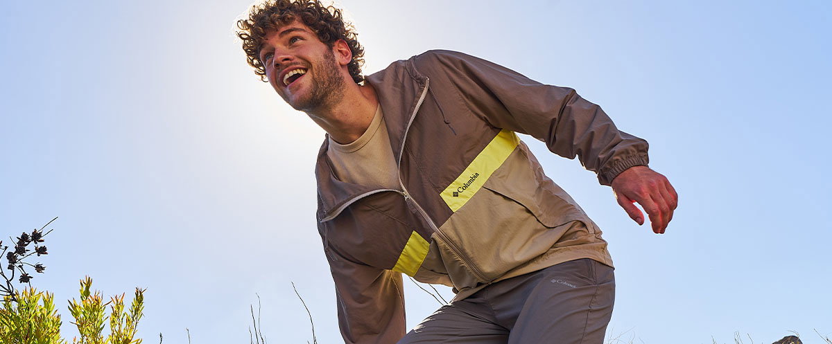 A person in a lightweight brown jacket with a bright panel leans forward outdoors under a clear sky, surrounded by low vegetation.