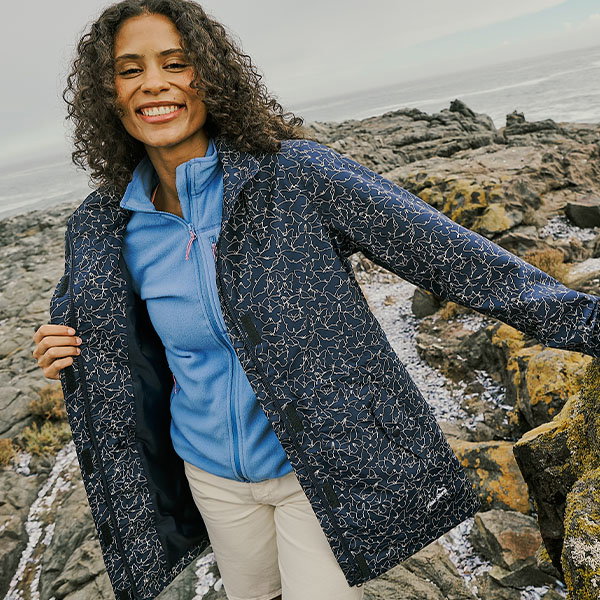 A person in a patterned jacket stands on a rocky coastline, holding the jacket open to reveal a blue layer underneath, with the sea and rugged rocks in the background.