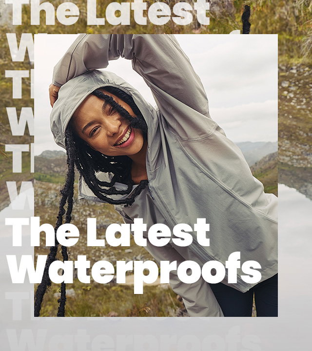 A person in a light grey hooded waterproof jacket leans sideways in a rocky, mountainous landscape with grasses and overcast skies, while large white text across the image reads “The Latest Waterproofs.”