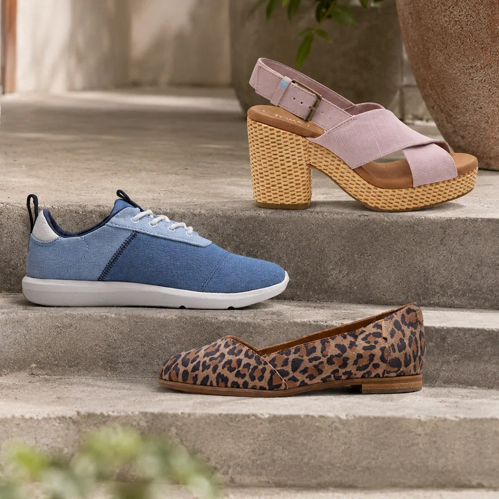 Three TOMS shoes displayed on steps: blue canvas sneaker, pink woven platform sandal, and leopard print loafer.