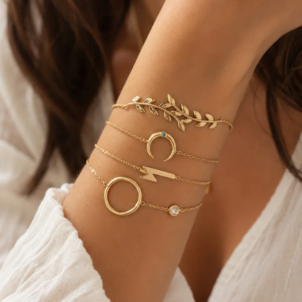 Gold charm bracelets stacked on wrist featuring leaf, crescent, lightning bolt, and circle designs with gemstones