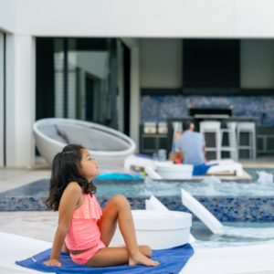 A young girl in a bright swimsuit sits on a blue towel beside a contemporary outdoor pool. She faces away from the camera, looking toward a man seated near an outdoor kitchen or bar area. The setting features sleek white lounge furniture and a mosaic-tiled pool and wall. The scene suggests a relaxed, family-oriented day in a modern backyard or resort environment.