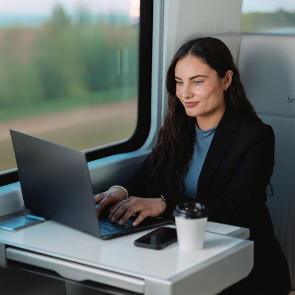 A woman in business attire works on a laptop while seated at a table on a modern train. She faces the window, with blurred greenery visible outside, suggesting the train is in motion. A smartphone and a takeaway coffee cup rest on the table beside her computer. The scene conveys productivity, remote work, and travel in a comfortable, contemporary setting.
