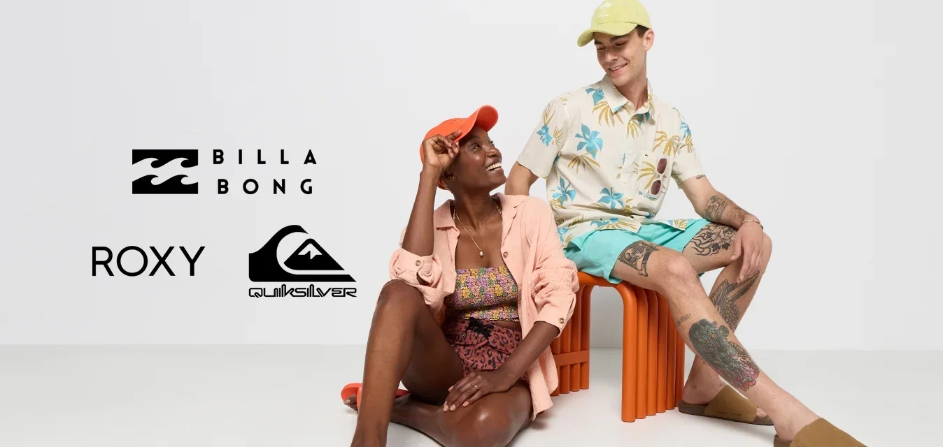 Twee personen zitten in een minimalistische omgeving. De één draagt een oranje pet, een lichtroze shirt en een gebloemd badpak; de ander heeft een lichtgroene pet, een bloemenhemd en shorts aan. Beiden dragen instapsandalen.
