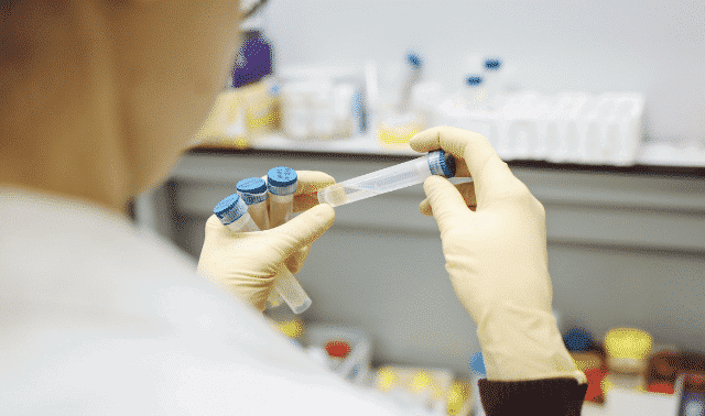 A person wearing gloves is holding up a test tube filled with a yellow liquid while other test tubes and laboratory materials are visible in the background.