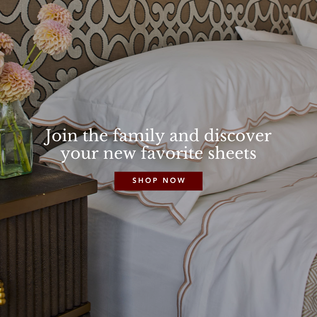 A bed with white sheets and pillows featuring a scalloped taupe trim, next to a nightstand with a vase of pink flowers.