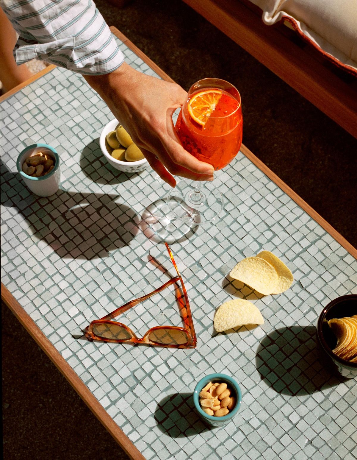 Hand reaching for an orange cocktail with citrus slice on a mosaic table with sunglasses, bowls of olives, nuts and crisps