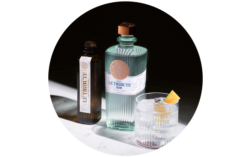 Le Tribute ribbed green gin bottle, small brown bottle and glass tumbler with gin, ice and lemon peel on marble