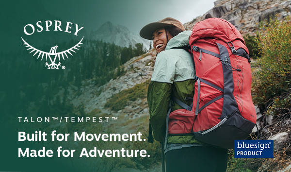 Osprey Talon & Tempest: Built for movement, made for adventure. A woman walks on a mountain trail, laughing, an Osprey Tempest 33L Daypack on her back.
