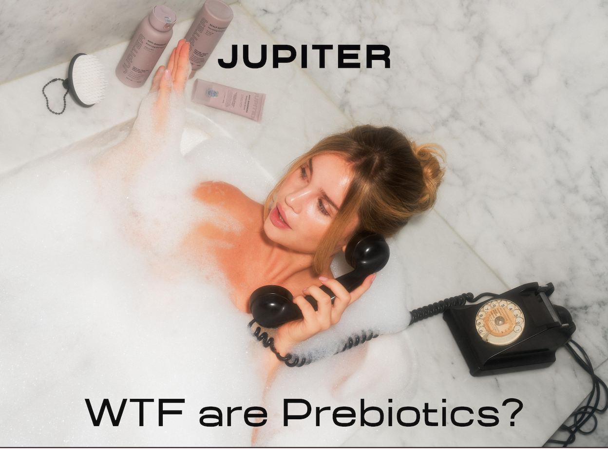 Woman relaxing in a bubble bath holding a vintage rotary phone receiver, with Jupiter Anti-Dandruff Balancing Shampoo, Scalp Soothing Conditioner, Anti-Dandruff Restoring Serum, and a Jupiter scalp brush arranged on the tub edge. Headline reads "WTF are Prebiotics?"