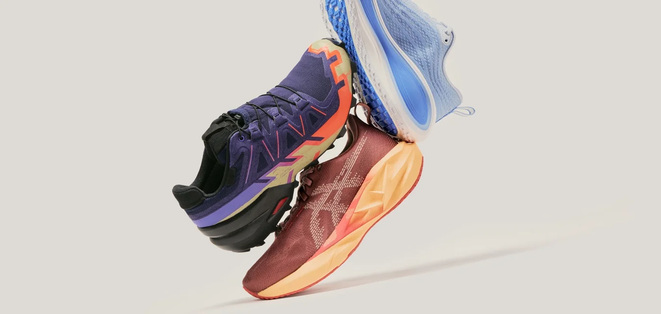 Drei bunte Sportschuhe, die in der Luft übereinander gestapelt sind, auf einem schlichten hellgrauen Hintergrund. Die Schuhe sind in Blau, Lila und rot-orange gestaltet.