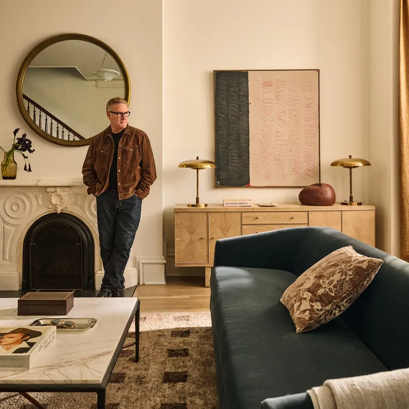  Todd Snyder leaning against a fireplace in a living room full of his furniture in collaboration with CB2. 