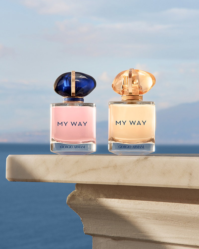 Two My Way perfume bottles with iconic stone-shaped caps on a marble ledge overlooking a calm sea and distant mountains.