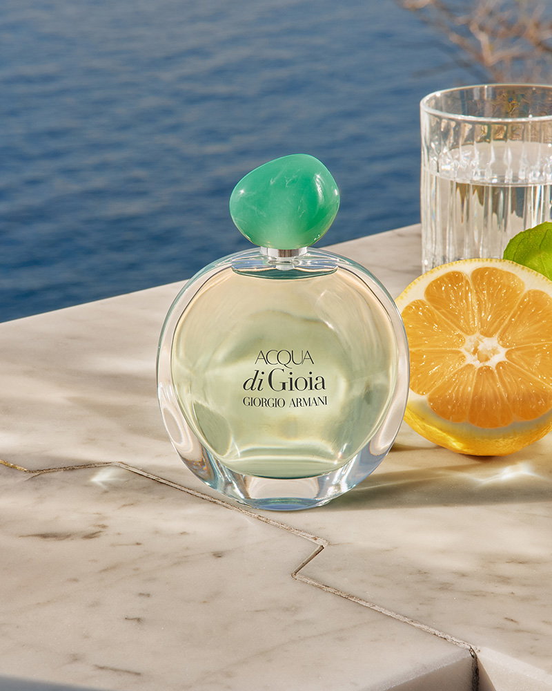 A bottle of Acqua di Gioia perfume with a green cap on a marble surface next to a lemon slice and a glass of water, overlooking the sea.