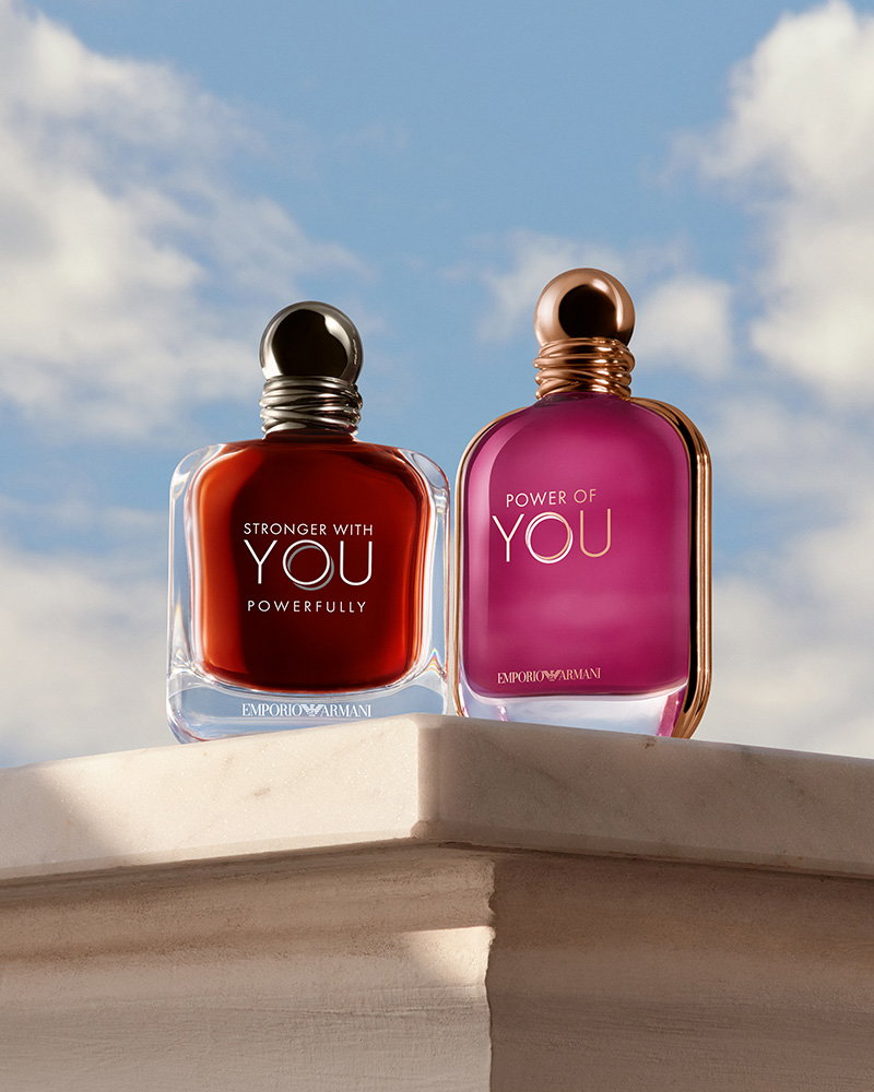 Two Armani perfume bottles, Stronger With You Powerfully and Power of You, placed on a marble ledge against a bright blue sky with white clouds.
