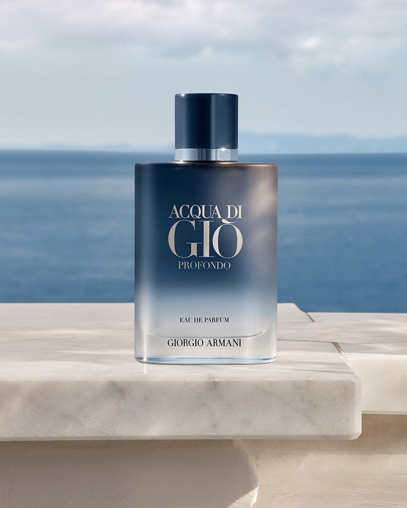A bottle of Acqua di Giò Profondo perfume with a blue gradient on a marble ledge with the ocean in the background.