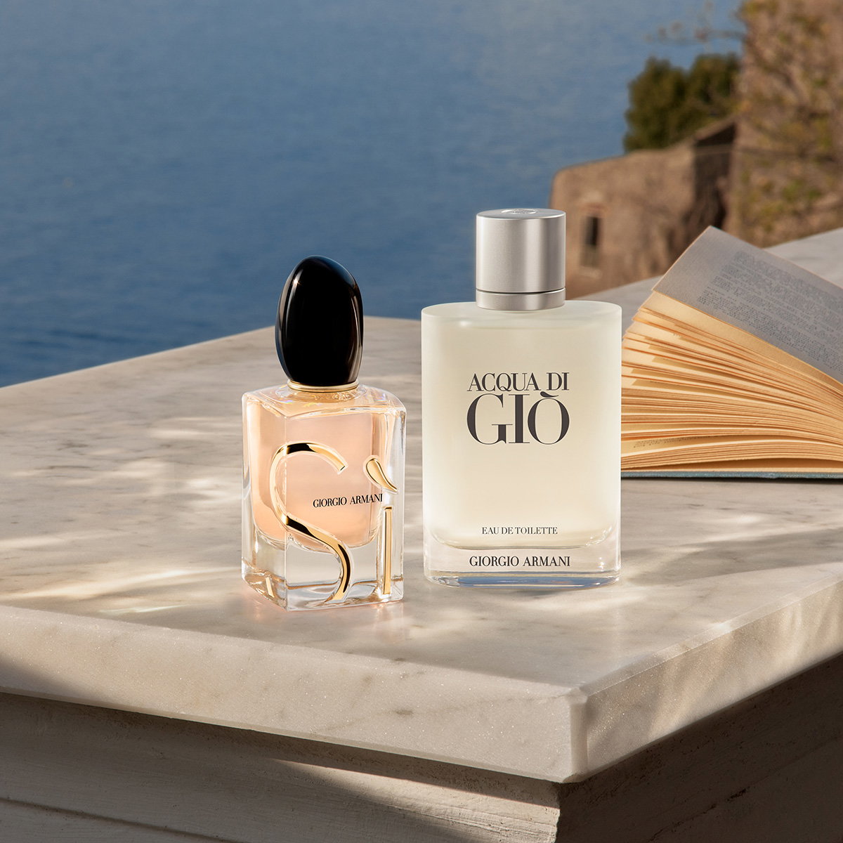 Two Armani perfume bottles, Sì and Acqua di Giò, sitting on a marble balcony next to an open book with the Mediterranean coast in the background.