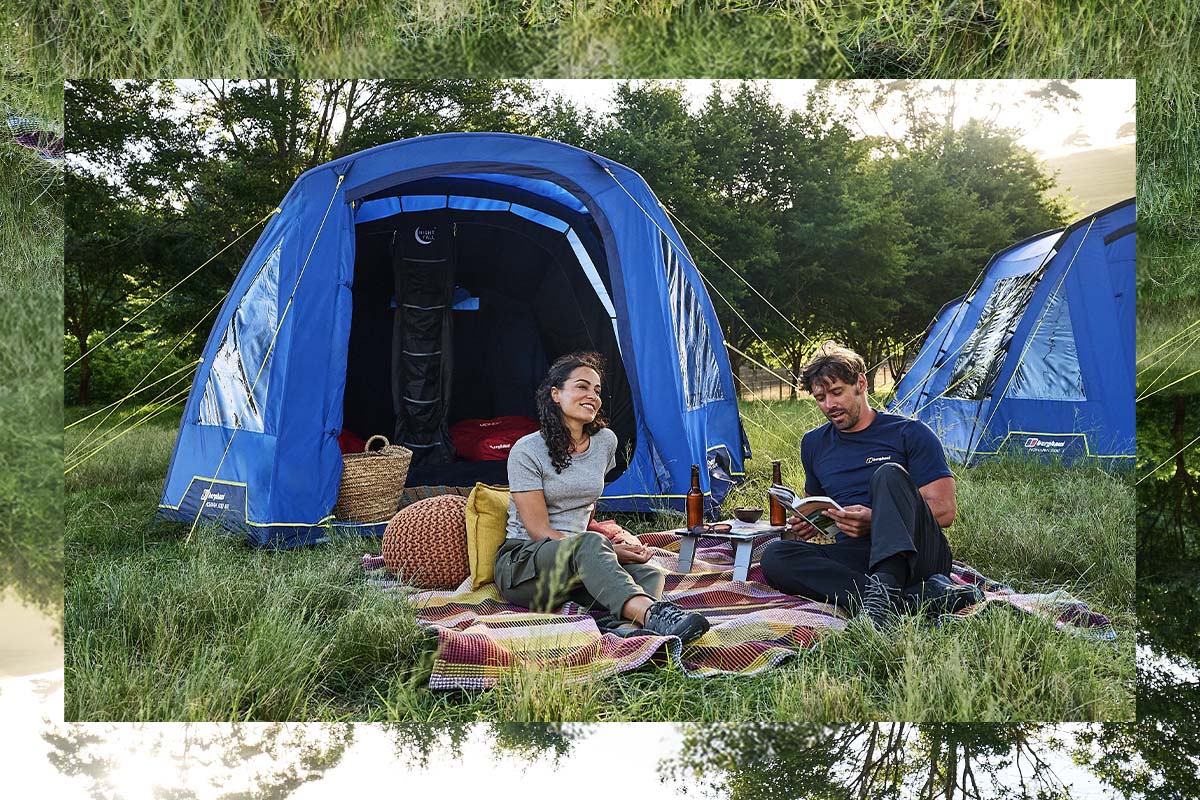 Two people sit on a picnic blanket in a grassy campsite beside blue tents, with trees in the background and camping gear arranged nearby.