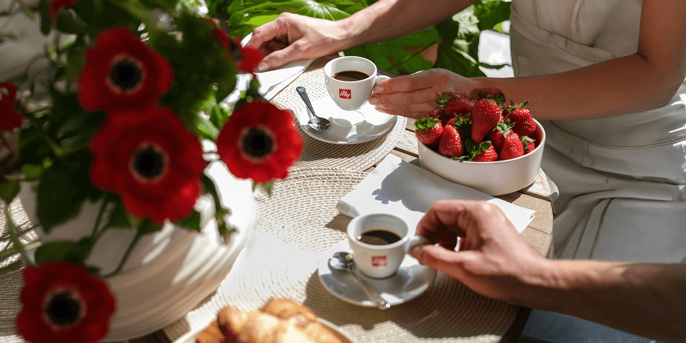 Two people enjoy espresso and fresh strawberries at a sunlit outdoor table