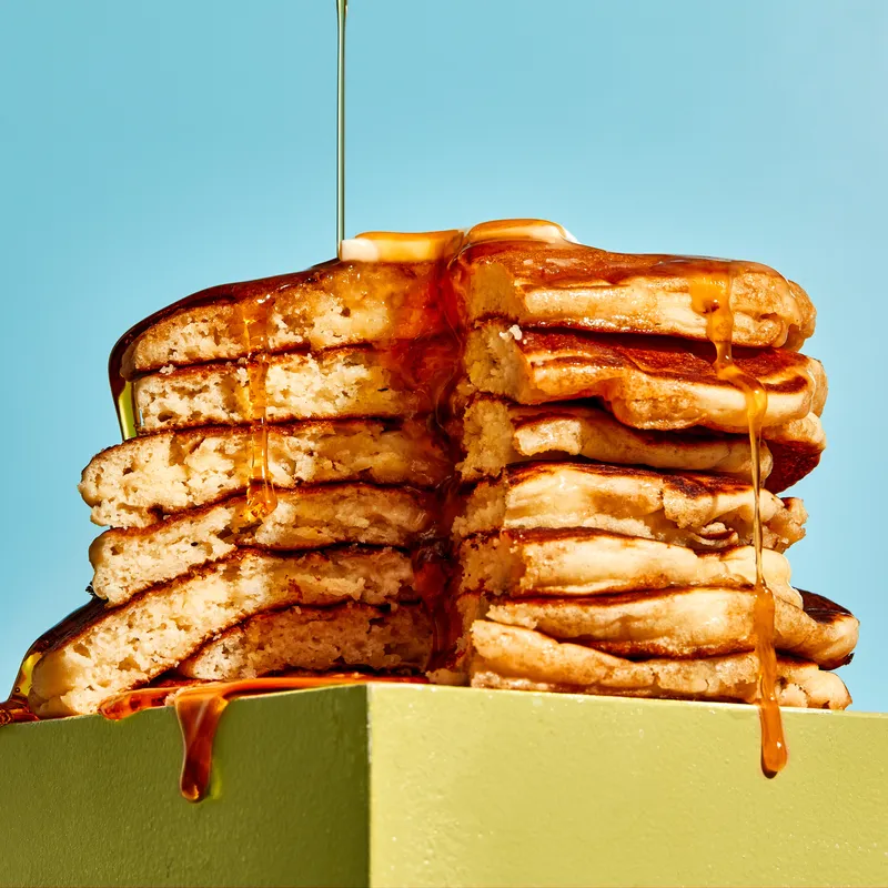 A stack of seven fluffy pancakes on table top, topped with butter, with syrup drizzling over the sides from above.