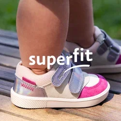Kleinkind trägt lila, pinke und weiße Sneakers und steht auf einem hölzernen Deck im Freien, mit verschwommenem Gras im Hintergrund.