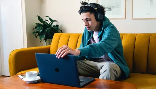 Person sitting in a yellow couch with headphones on while working on a laptop