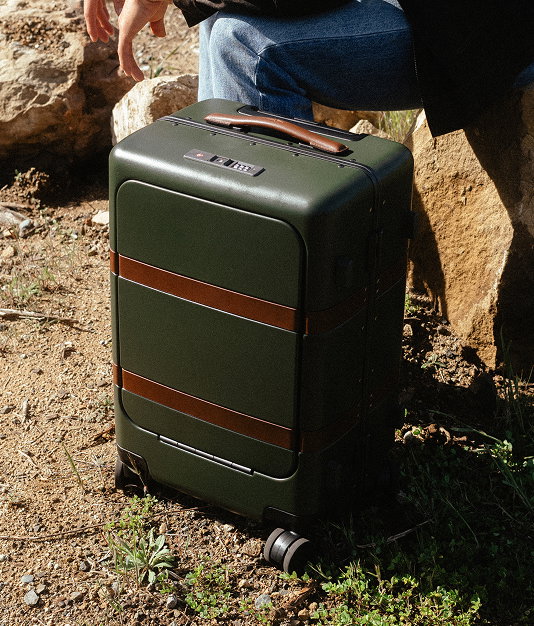 Green hardshell carry-on suitcase with brown leather straps on a rocky trail