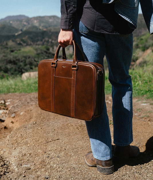 Man relaxing on an outdoor bench with a brown leather briefcase beside him