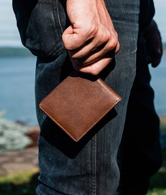 Brown leather bifold wallet being held outside in front of a man wearing denim