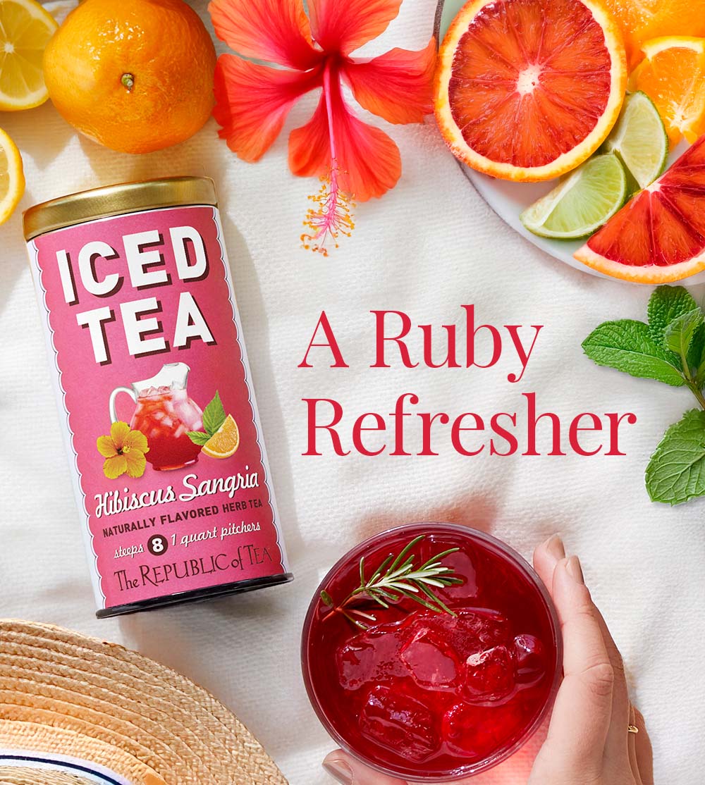 Hibiscus Sangria Iced Tea tin on a beach blanket surrounded by citrus fruits, hibiscus flower, mint leaves and a beach hat at the bottom corner with a persons hand holding an iced tea glass.