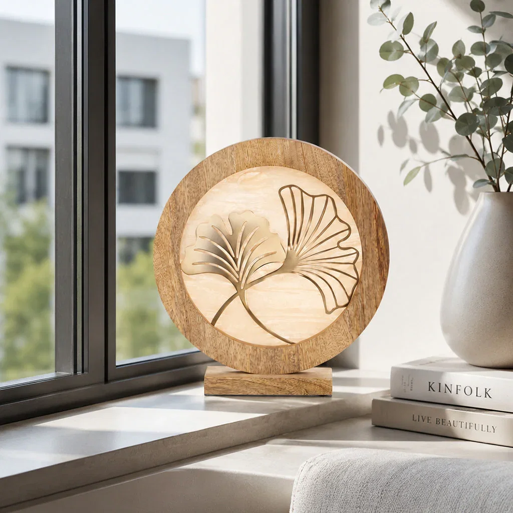 Wooden decorative disc with carved ginkgo leaf design on windowsill with books and potted plant.