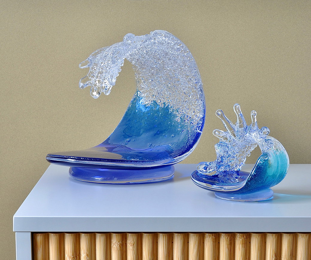 Two vibrant blue glass sculptures depicting cresting ocean waves, one small and one large, displayed on a sideboard. - Shop Now.