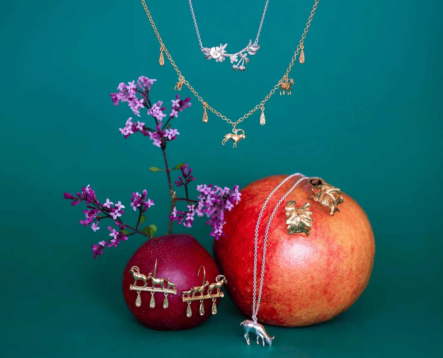 Gold and silver Summer Archive Sale jewellery styled with pomegranate, apple, and purple blooms against a teal background.