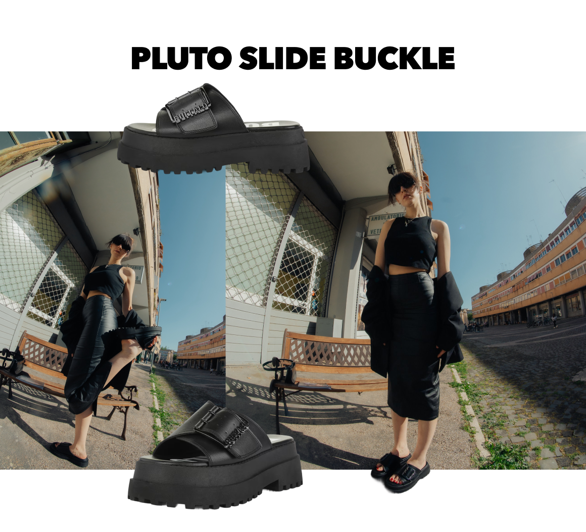 Black platform slide sandal with buckle detail, styled in an urban street scene with model wearing all-black outfit.